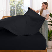 Woman putting sheet on a bed in a bright room with large windows.
