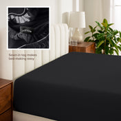 Fitted sheet on a bed with a close-up of a sewn-in tag, nightstand, lamp, and plant in a bedroom setting.