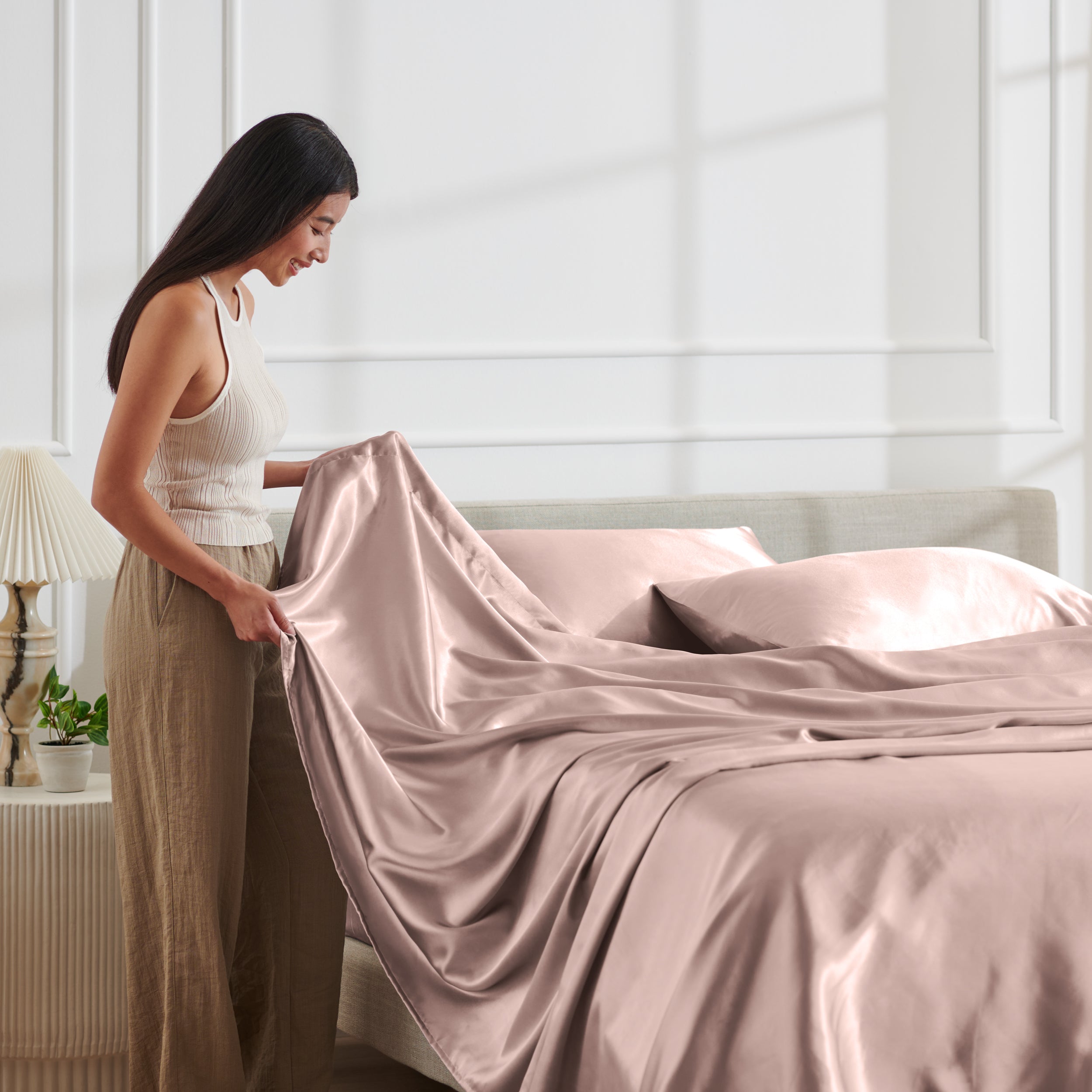 Woman arranging satin bedding on a bed in a bright bedroom.