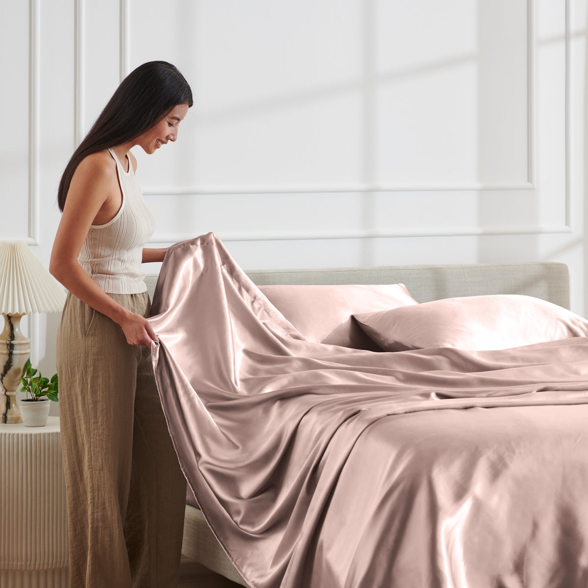 Woman arranging satin bedding on a bed in a bright bedroom.