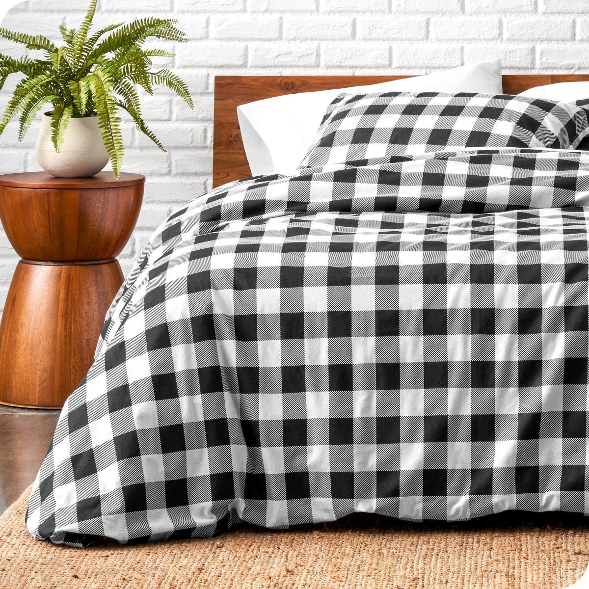 Bed with black and white checkered duvet cover next to a wooden side table with a plant, against a white brick wall.