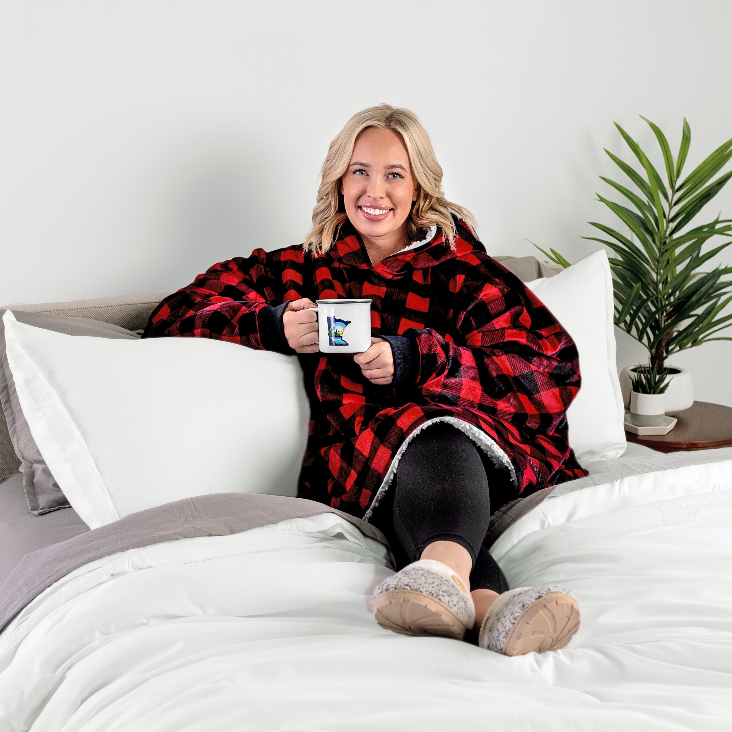 A woman sitting on a bed holding a mug wearing a sherpa blanket.
