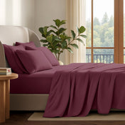 A neatly made bed with in a softly lit bedroom. A large green plant stands beside the bed, and a window with curtains is visible in the background.