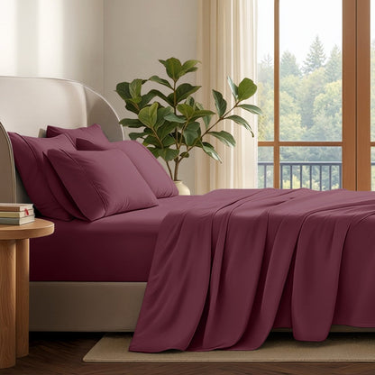 A neatly made bed with in a softly lit bedroom. A large green plant stands beside the bed, and a window with curtains is visible in the background.