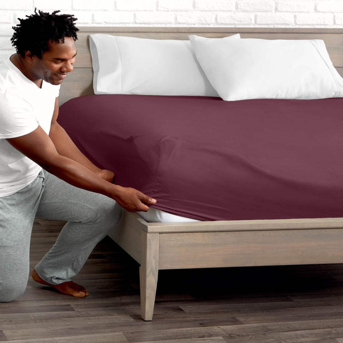 Man adjusting fitted sheet on a bed with white pillows.