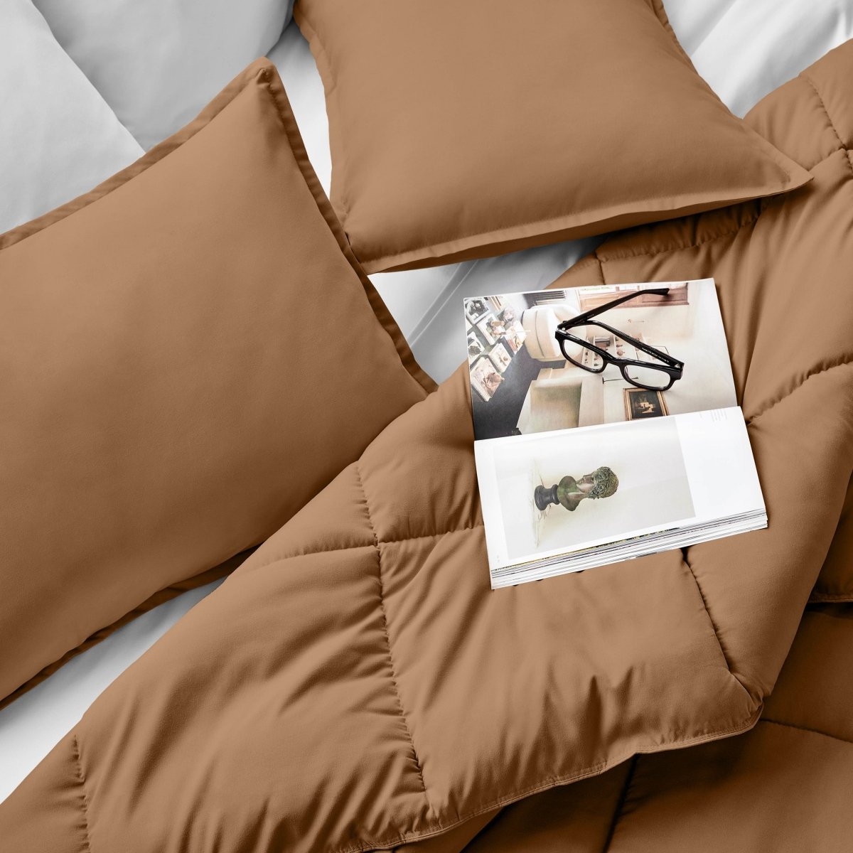 Comforter and pillows on a bed with a magazine and glasses.