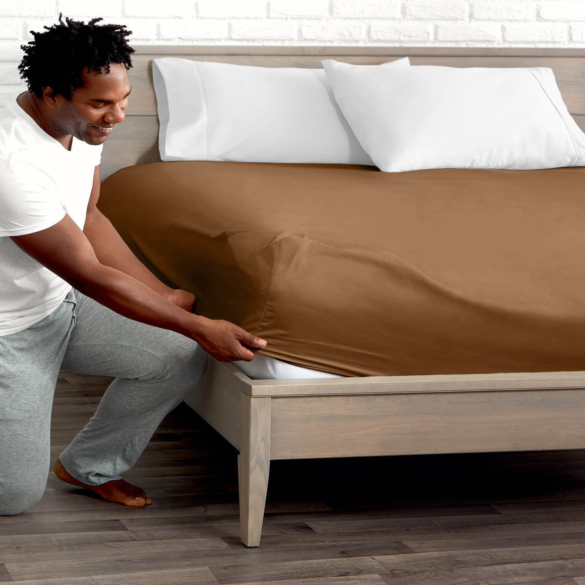 Man adjusting fitted sheet on a bed with white pillows.