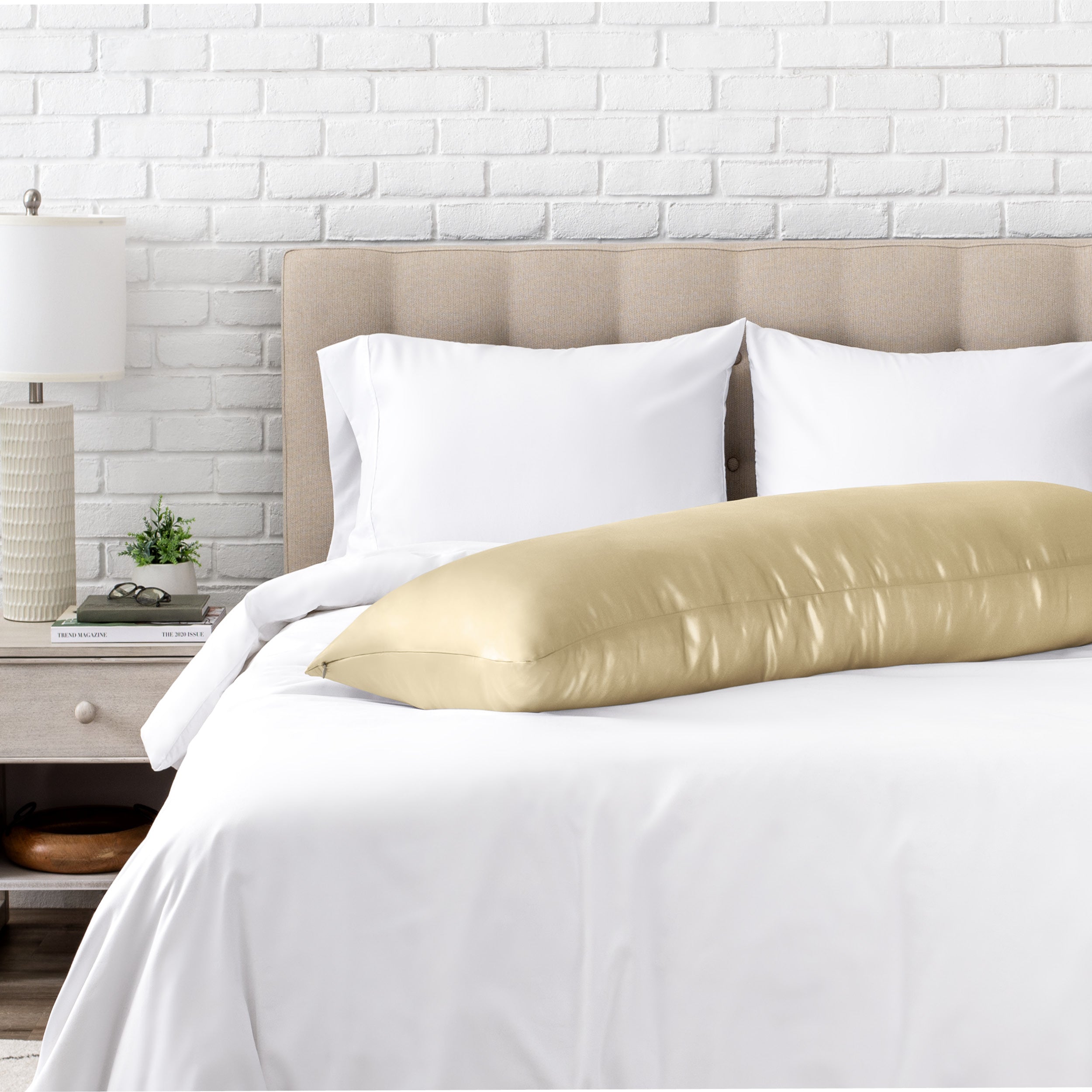 Bed with white bedding and a body pillow against a white brick wall.