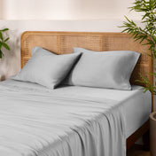 Bamboo bed sheets and pillows on a bed with a wicker headboard and plants in the background.