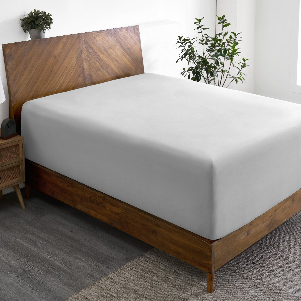 Wooden bed frame with a fitted sheet on the mattress in a room with plants and a nightstand.