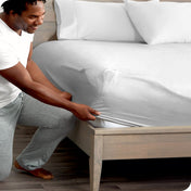 Man in white shirt and gray pants adjusting a fitted sheet on a bed.