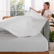 Woman putting sheet on a bed in a bright room with large windows.