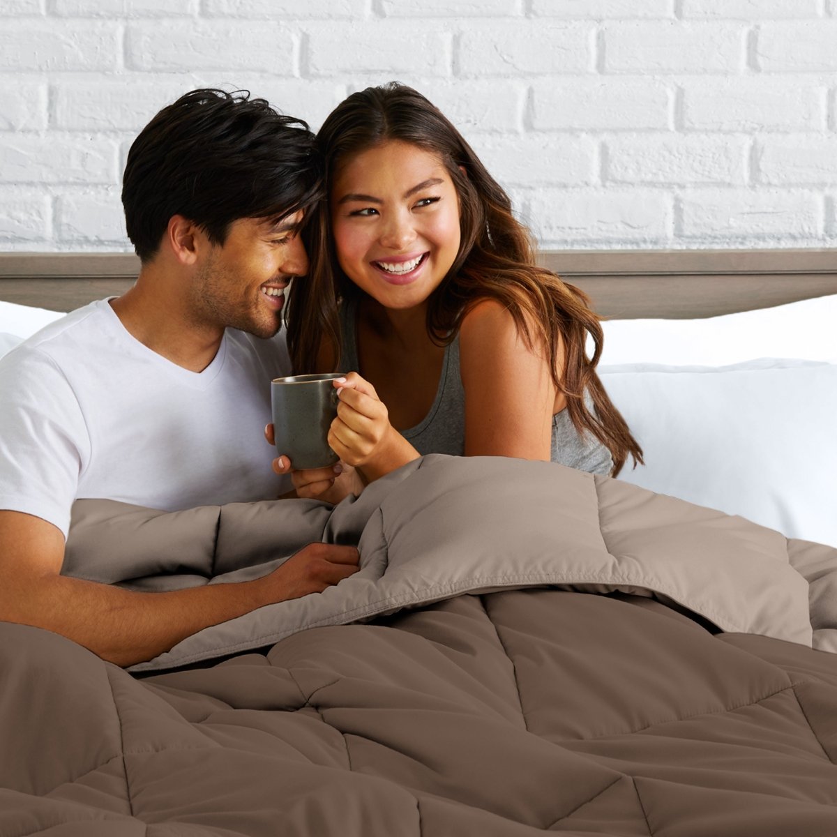 Man and woman sitting on a bed holding a mug, smiling at each other.
