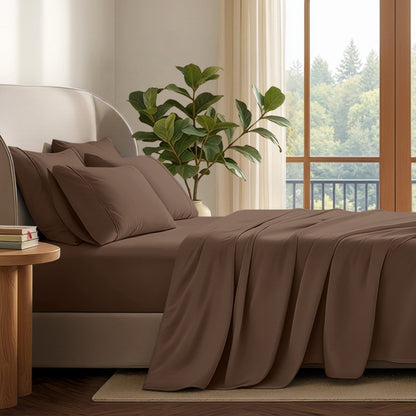 A neatly made bed with in a softly lit bedroom. A large green plant stands beside the bed, and a window with curtains is visible in the background.