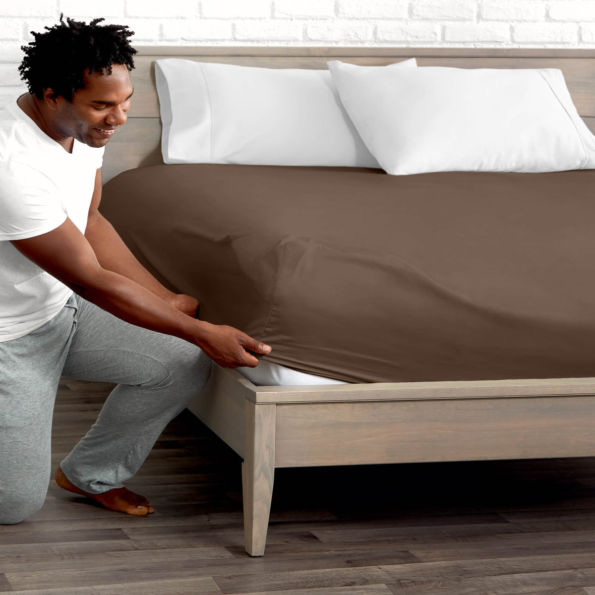 Man adjusting fitted sheet on a bed with white pillows.