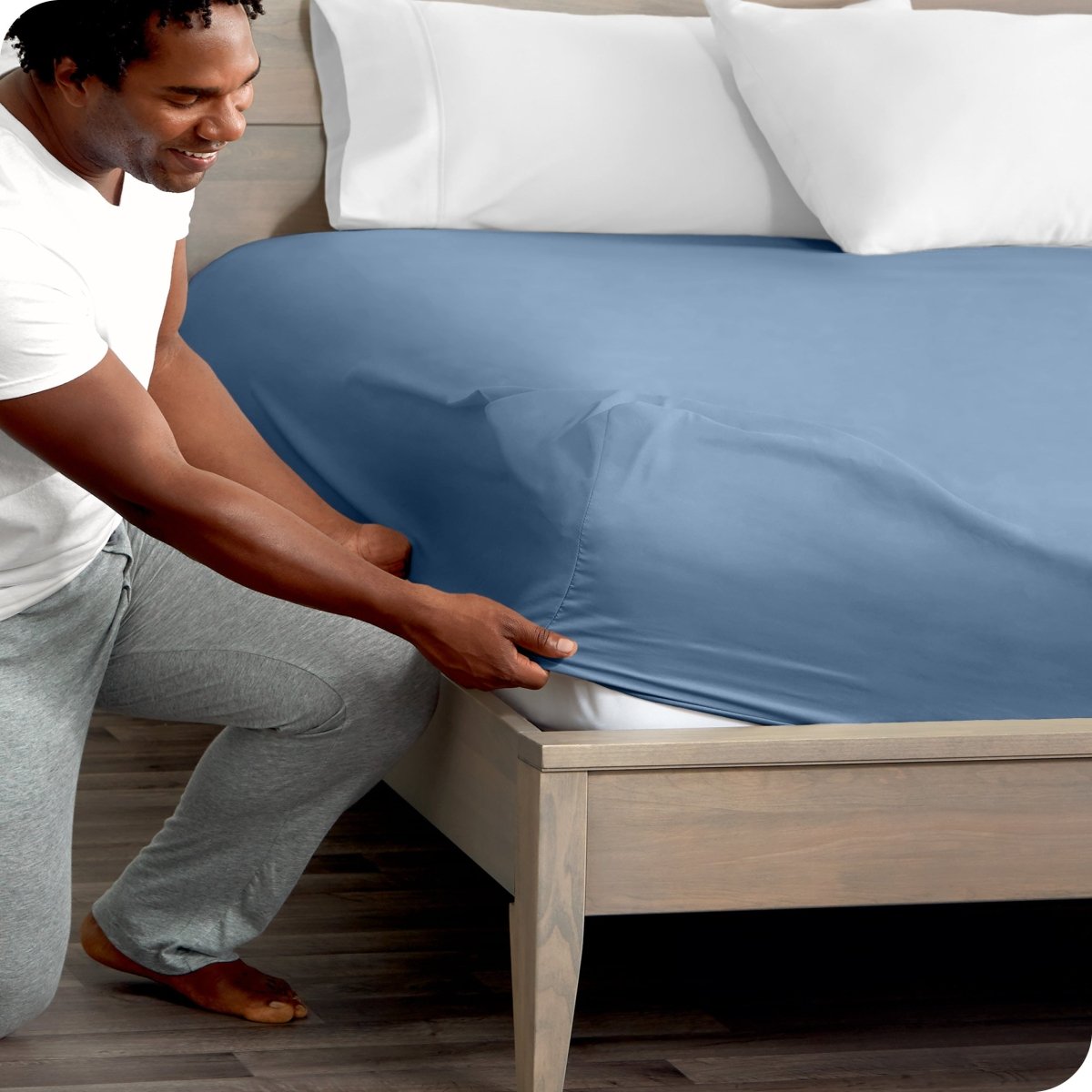 Man in white shirt and gray pants adjusting a fitted sheet on a bed.