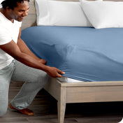Man in white shirt and gray pants adjusting a fitted sheet on a bed.