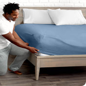 A man is kneeing and putting the corner of the fitted sheet on the mattress. The mattress is on a wooden bed frame.