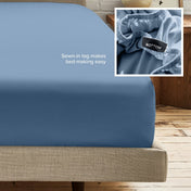A fitted bed sheet on a bed frame with a small inset close-up of the elastic and a sewn-in tag labeled BOTTOM. White text on the sheet reads, 'Sewn-in tag makes bed-making easy.