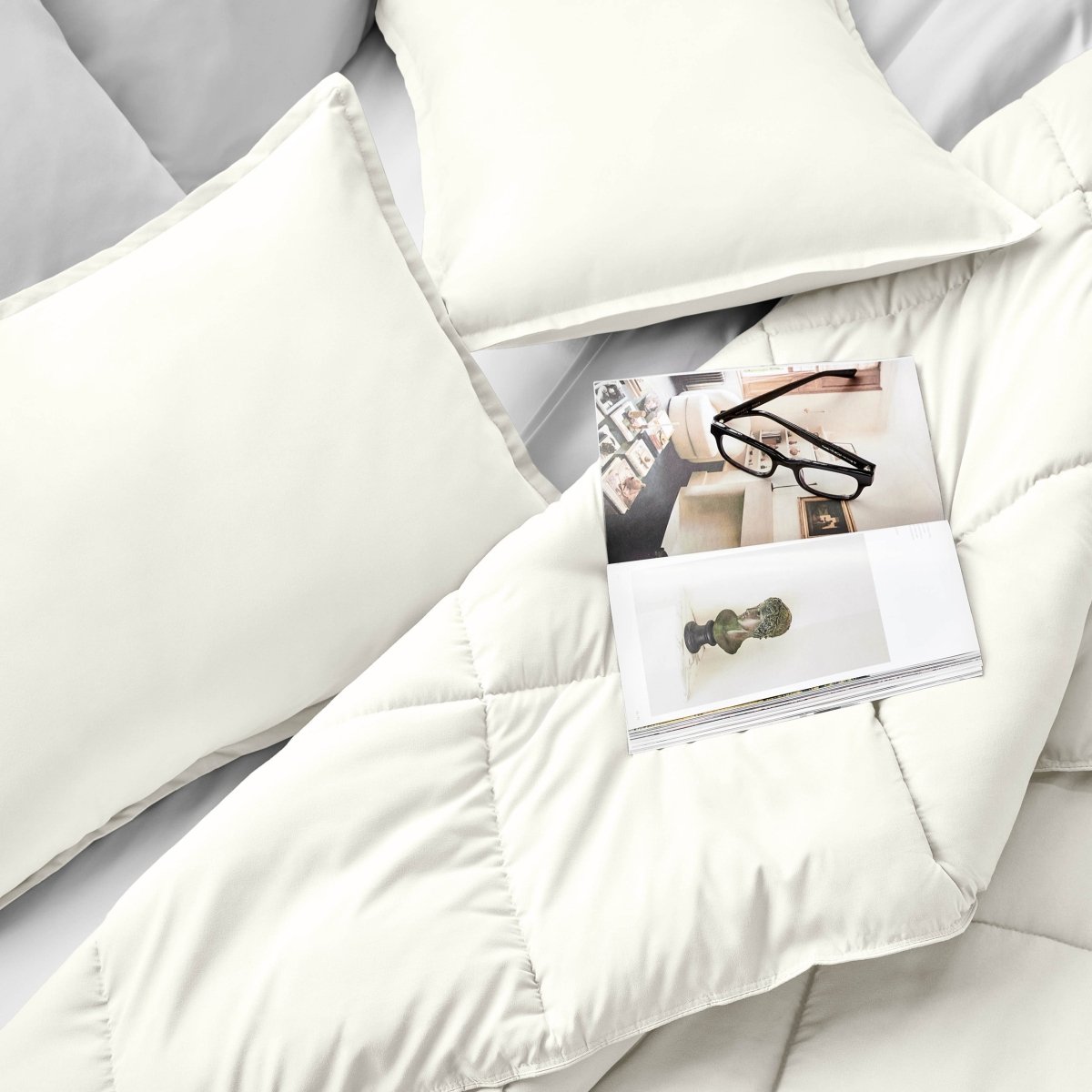 Comforter and pillows on a bed with a magazine and glasses.