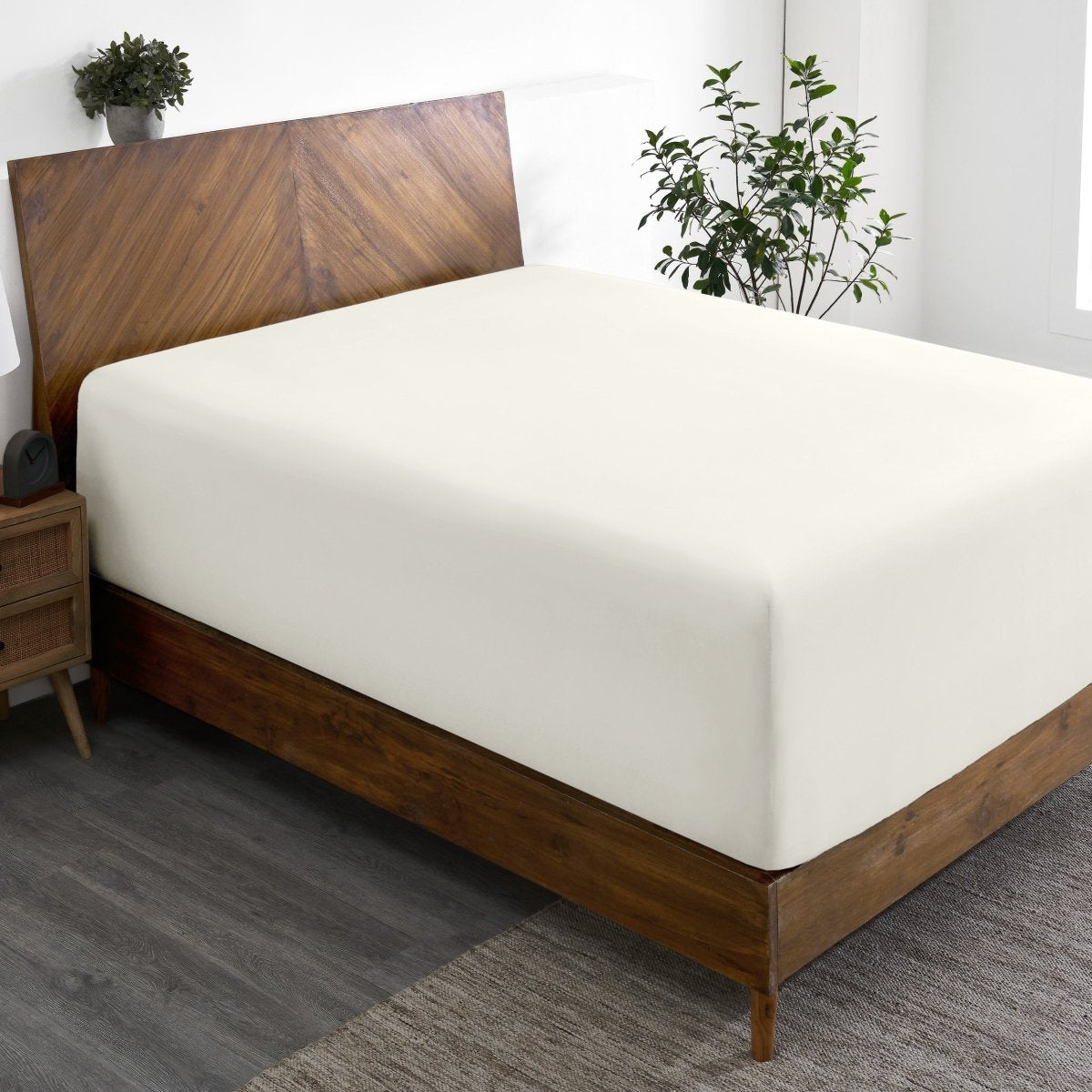 Wooden bed frame with a fitted sheet on the mattress in a room with plants and a nightstand.