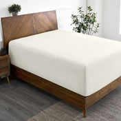 Wooden bed frame with a fitted sheet on the mattress in a room with plants and a nightstand.