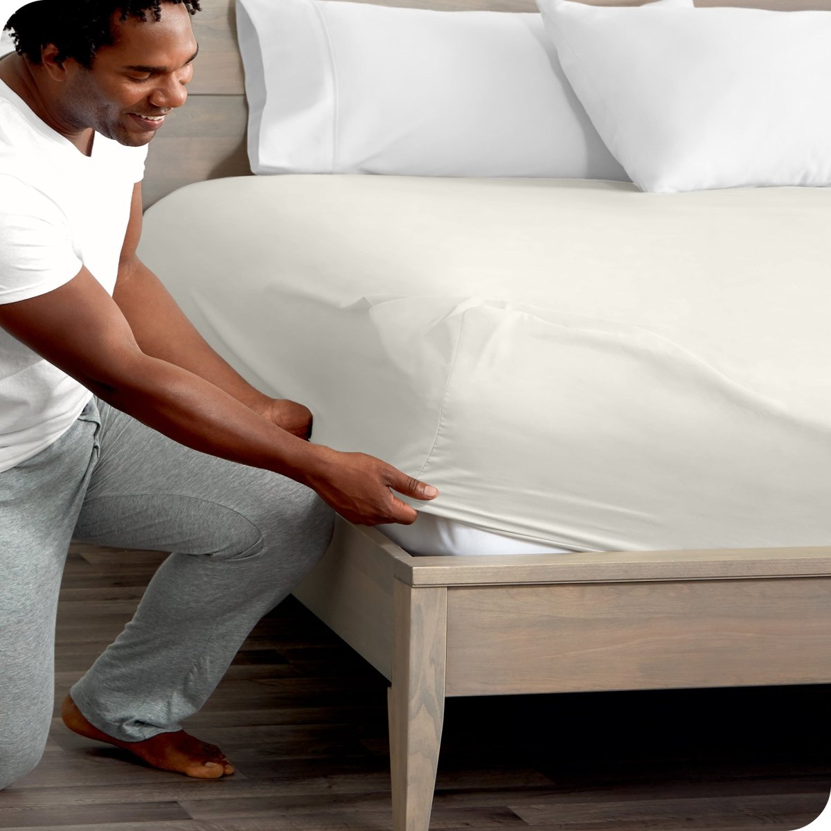 Man in white shirt and gray pants adjusting a fitted sheet on a bed.