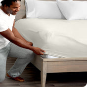 Man in white shirt and gray pants adjusting a fitted sheet on a bed.