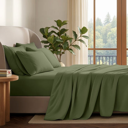 A neatly made bed with in a softly lit bedroom. A large green plant stands beside the bed, and a window with curtains is visible in the background.