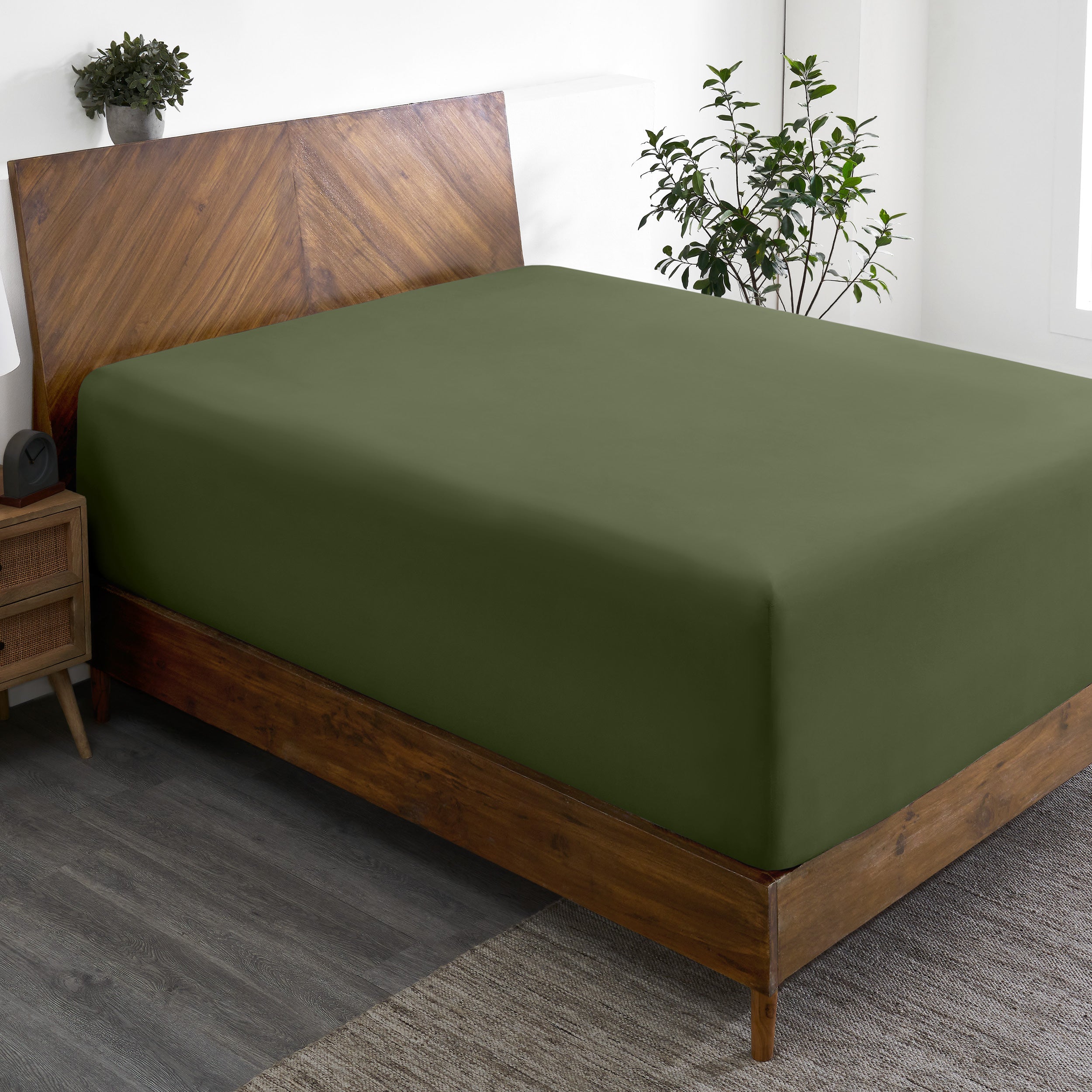 Wooden bed frame with a fitted sheet on the mattress in a room with plants and a nightstand.