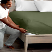Man in white shirt and gray pants adjusting a fitted sheet on a bed.