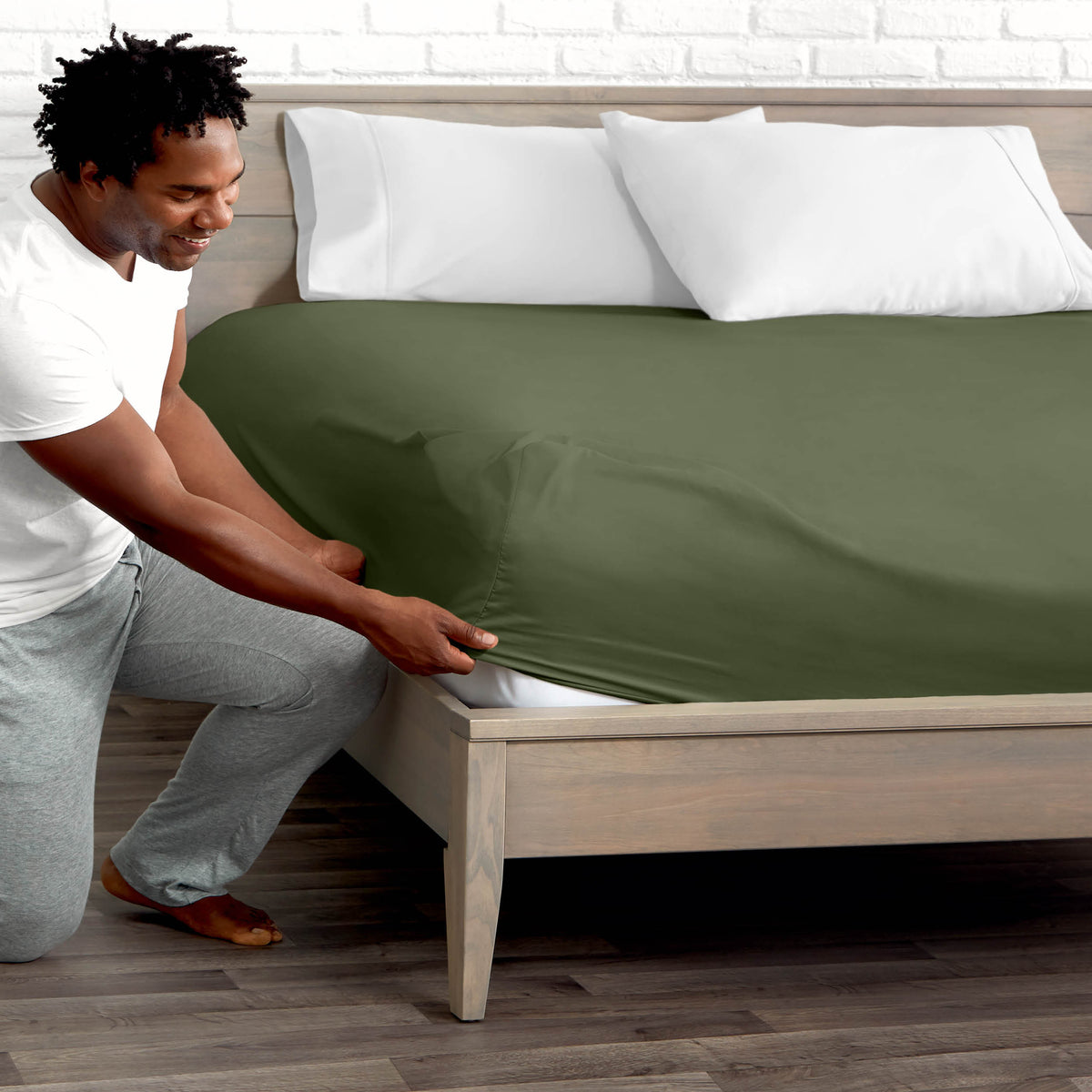 Man adjusting fitted sheet on a bed with white pillows.