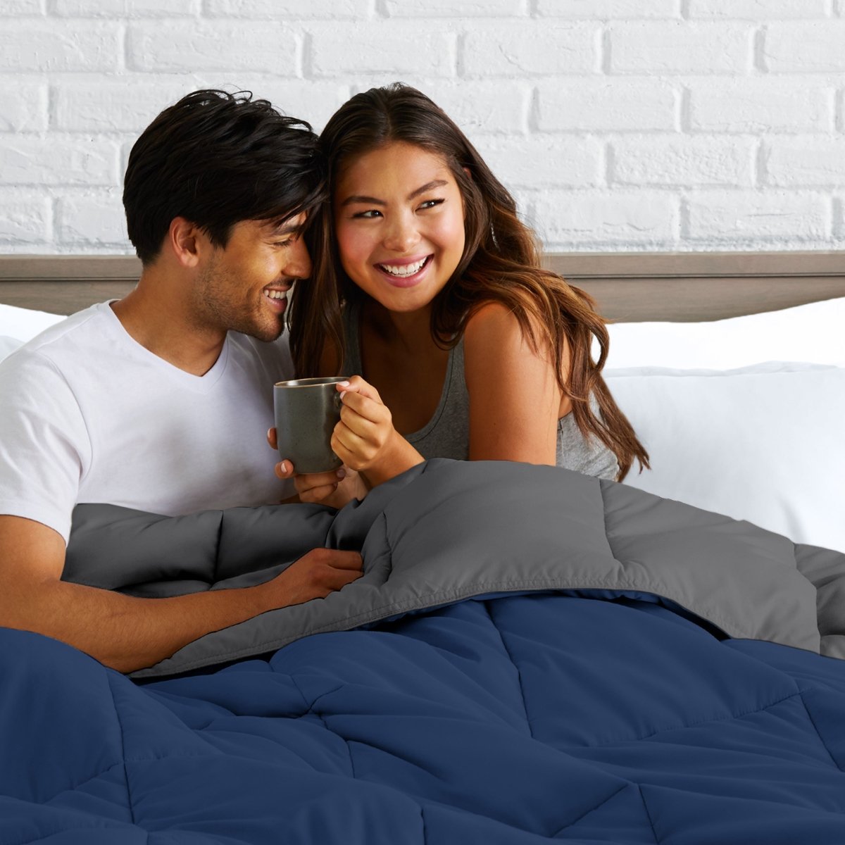 Man and woman sitting on a bed holding a mug, smiling at each other.