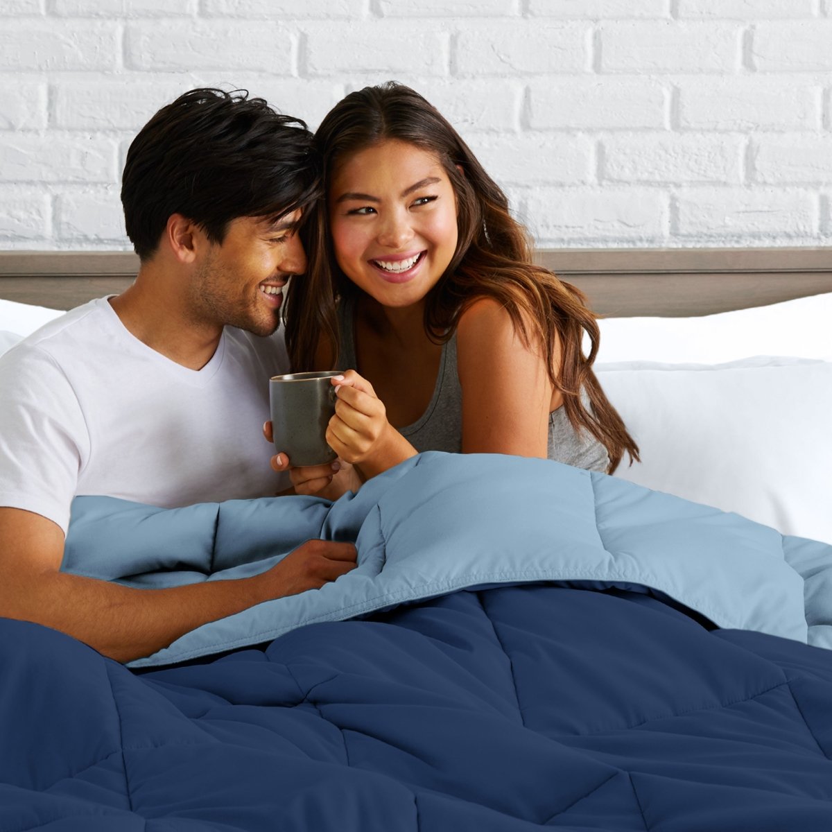 Man and woman sitting on a bed holding a mug, smiling at each other.