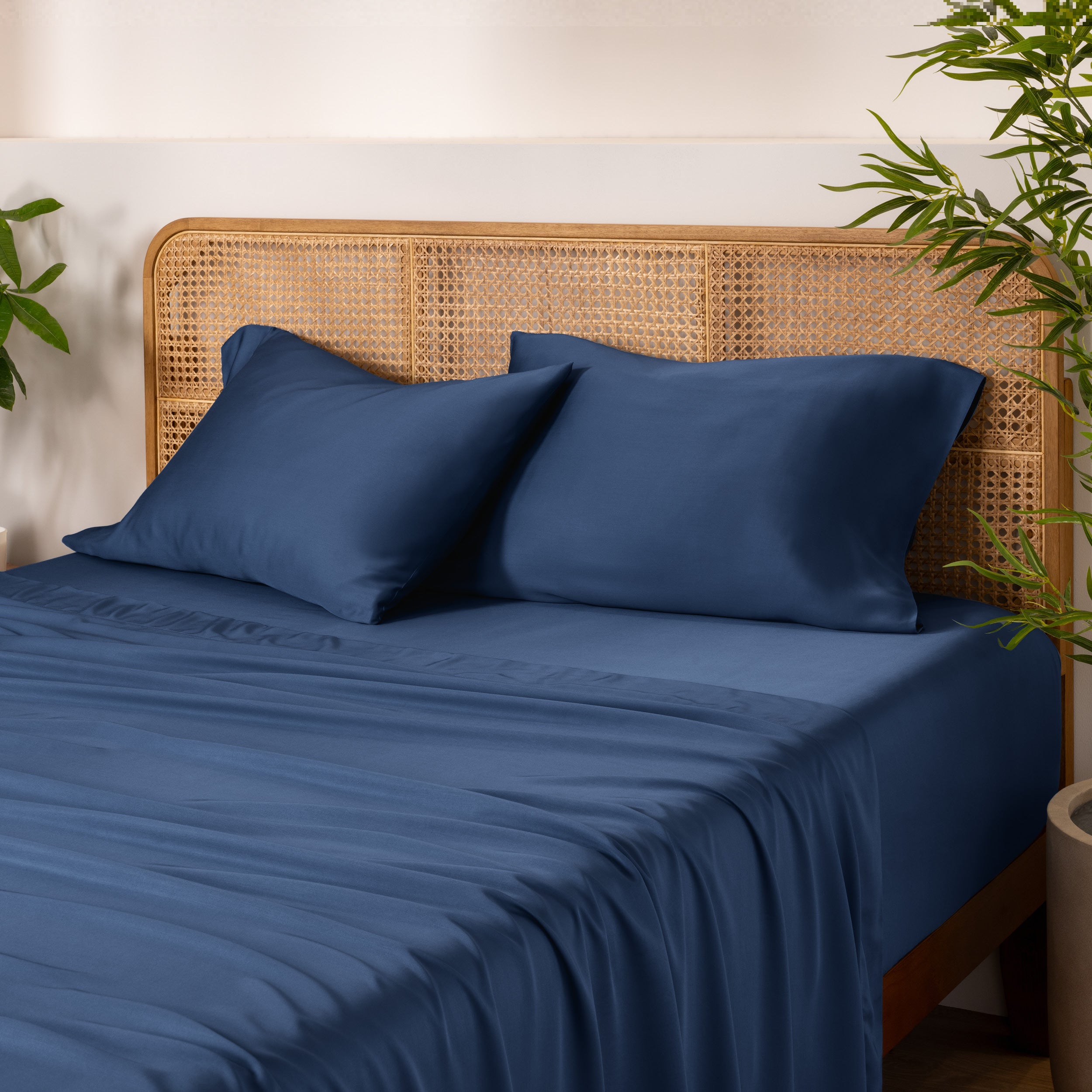 Bamboo bed sheets and pillows on a bed with a wicker headboard and plants in the background.