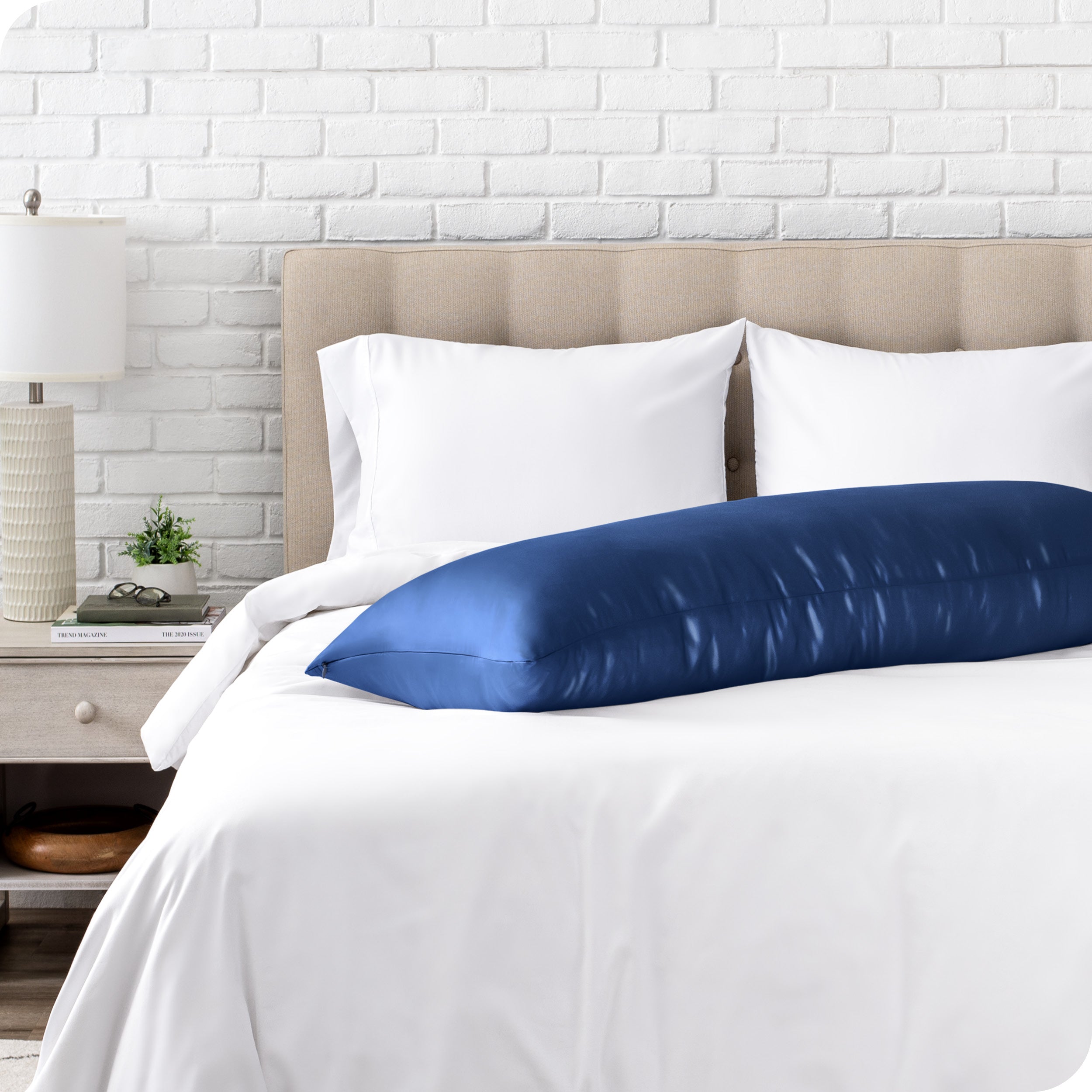 Bed with white bedding and a body pillow against a white brick wall.