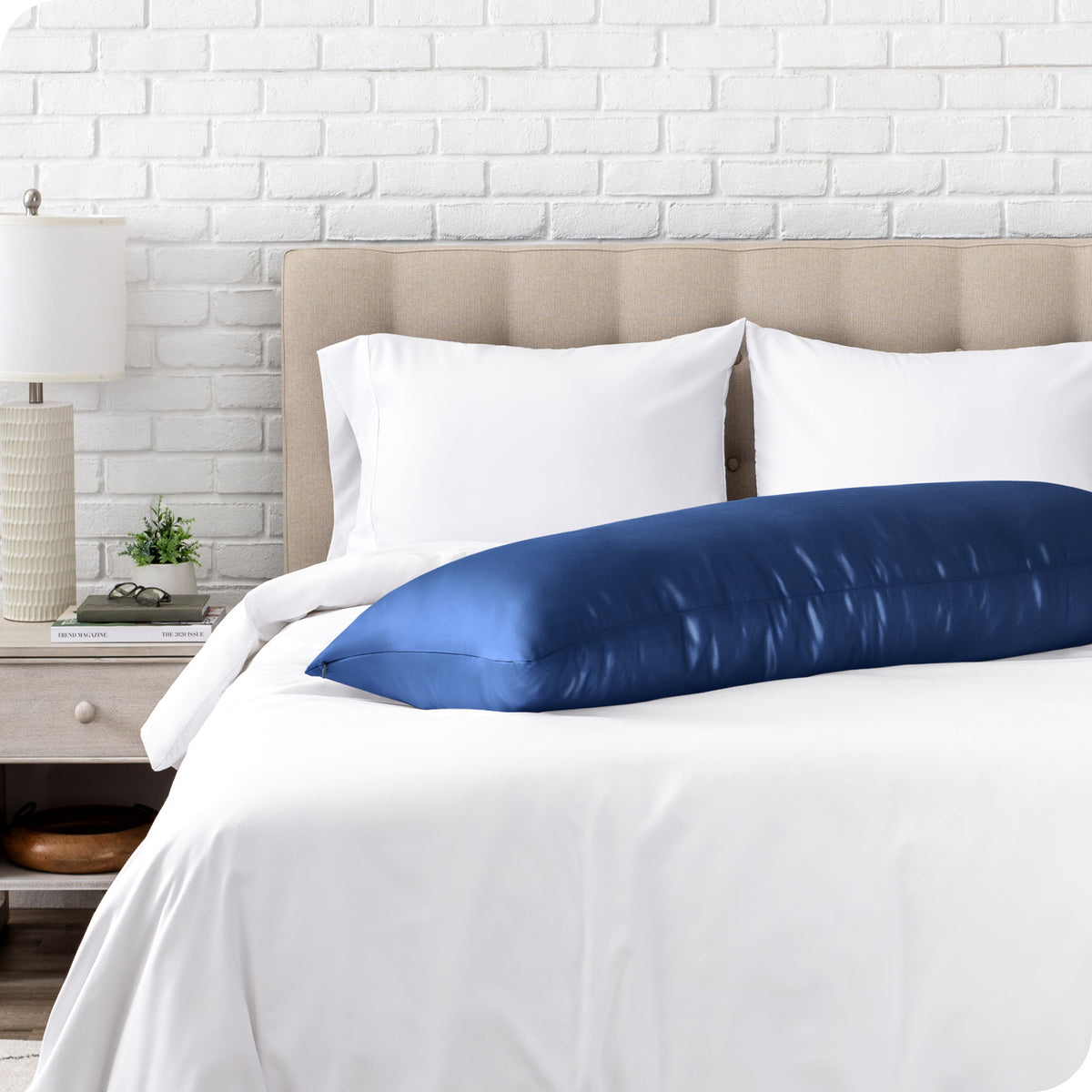 Bed with white bedding and a body pillow against a white brick wall.