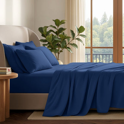 A neatly made bed with in a softly lit bedroom. A large green plant stands beside the bed, and a window with curtains is visible in the background.