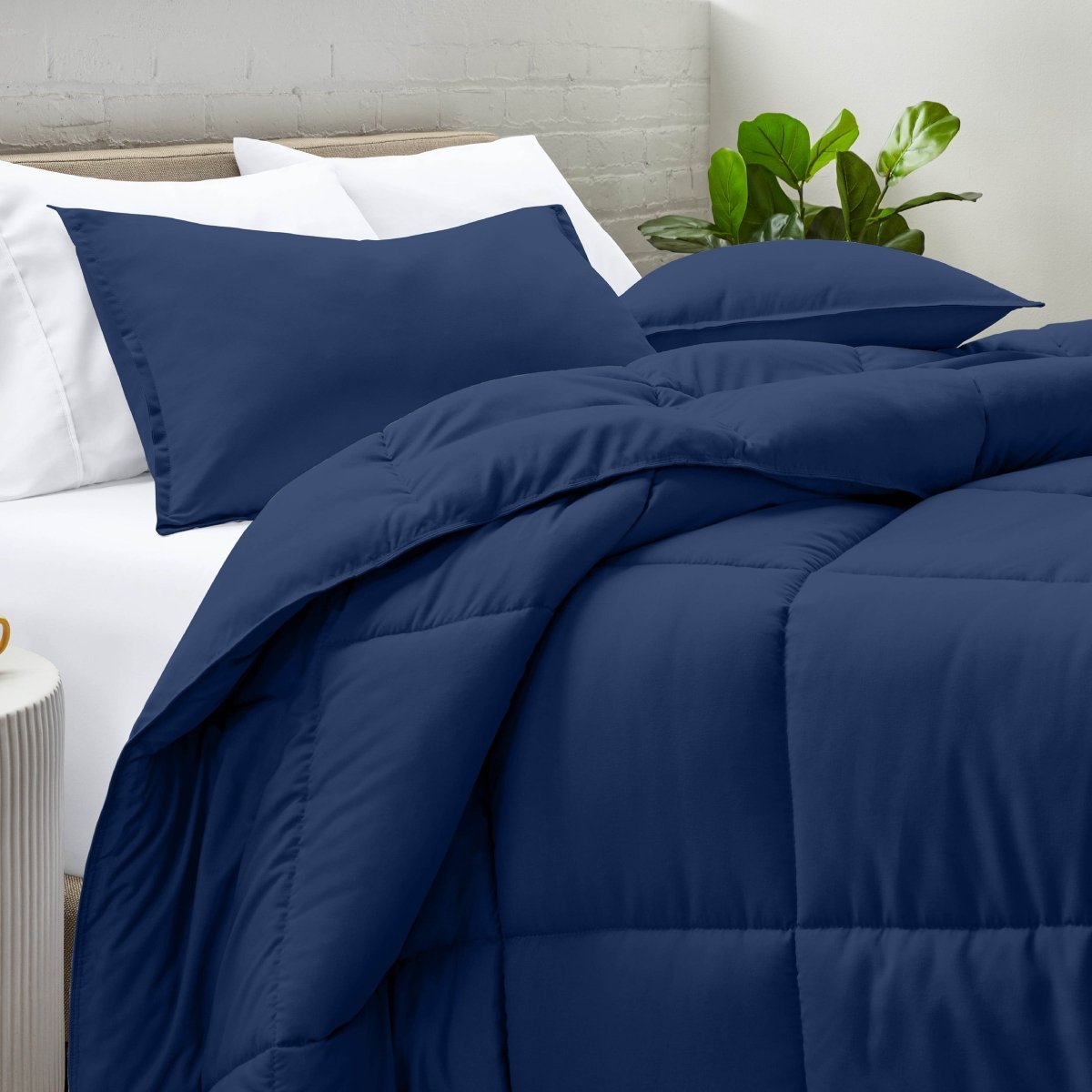 Comforter and pillow shams on a bed with a white brick wall and plant in the background