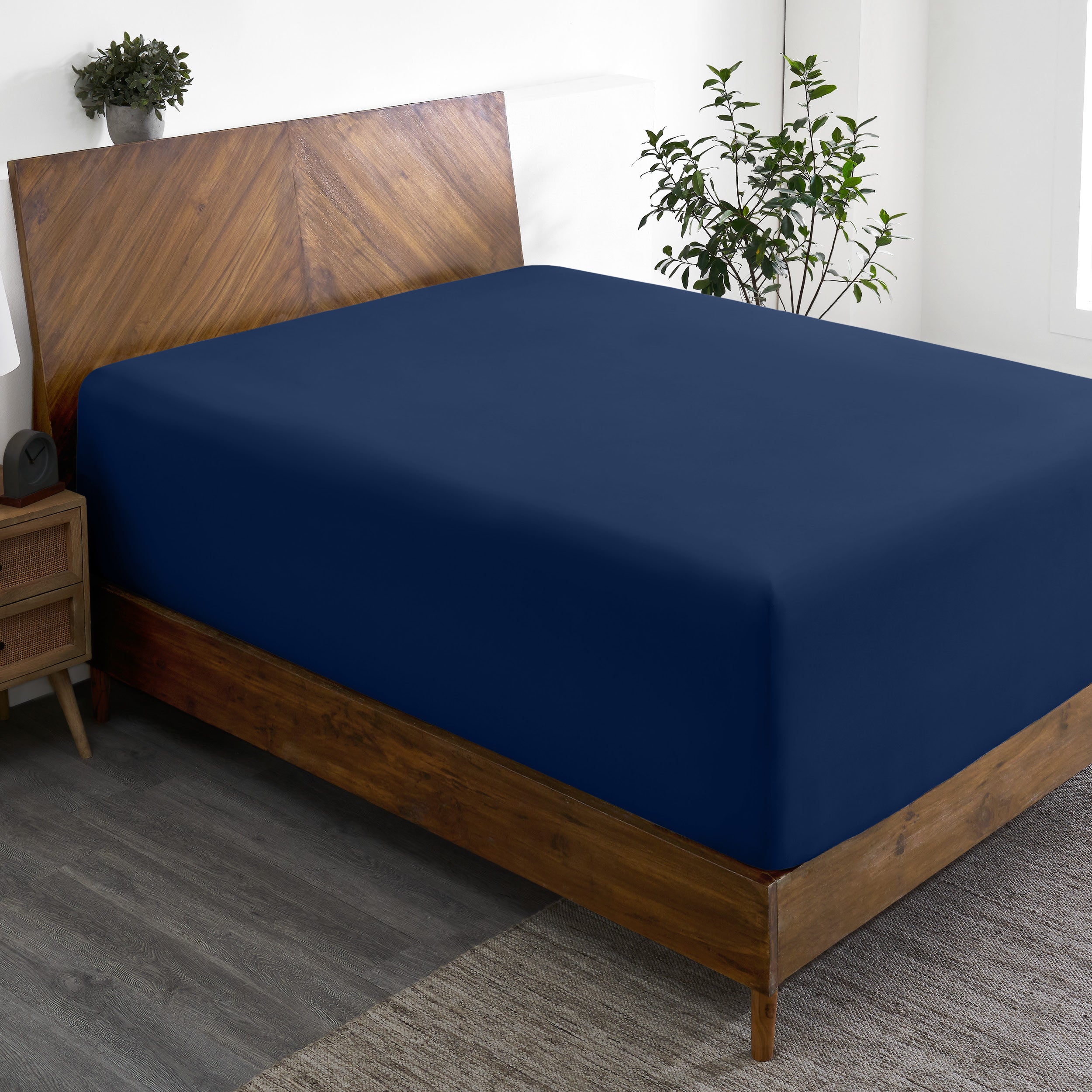 Wooden bed frame with a fitted sheet on the mattress in a room with plants and a nightstand.