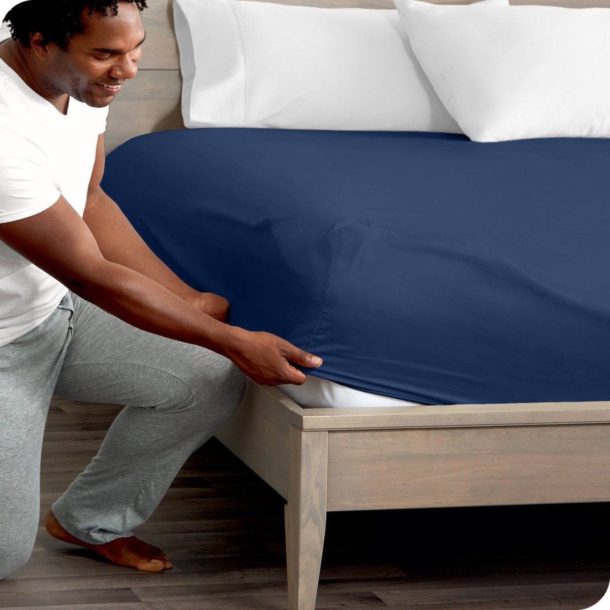 Man in white shirt and gray pants adjusting a fitted sheet on a bed.