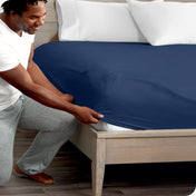 A man is kneeling while putting a fitted sheet on a bed