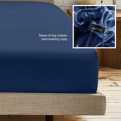 A fitted bed sheet on a bed frame with a small inset close-up of the elastic and a sewn-in tag labeled BOTTOM. White text on the sheet reads, 'Sewn-in tag makes bed-making easy.