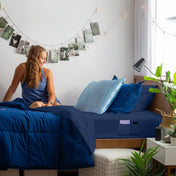 Woman sitting on a bed with blue bedding, surrounded by plants and decor.
