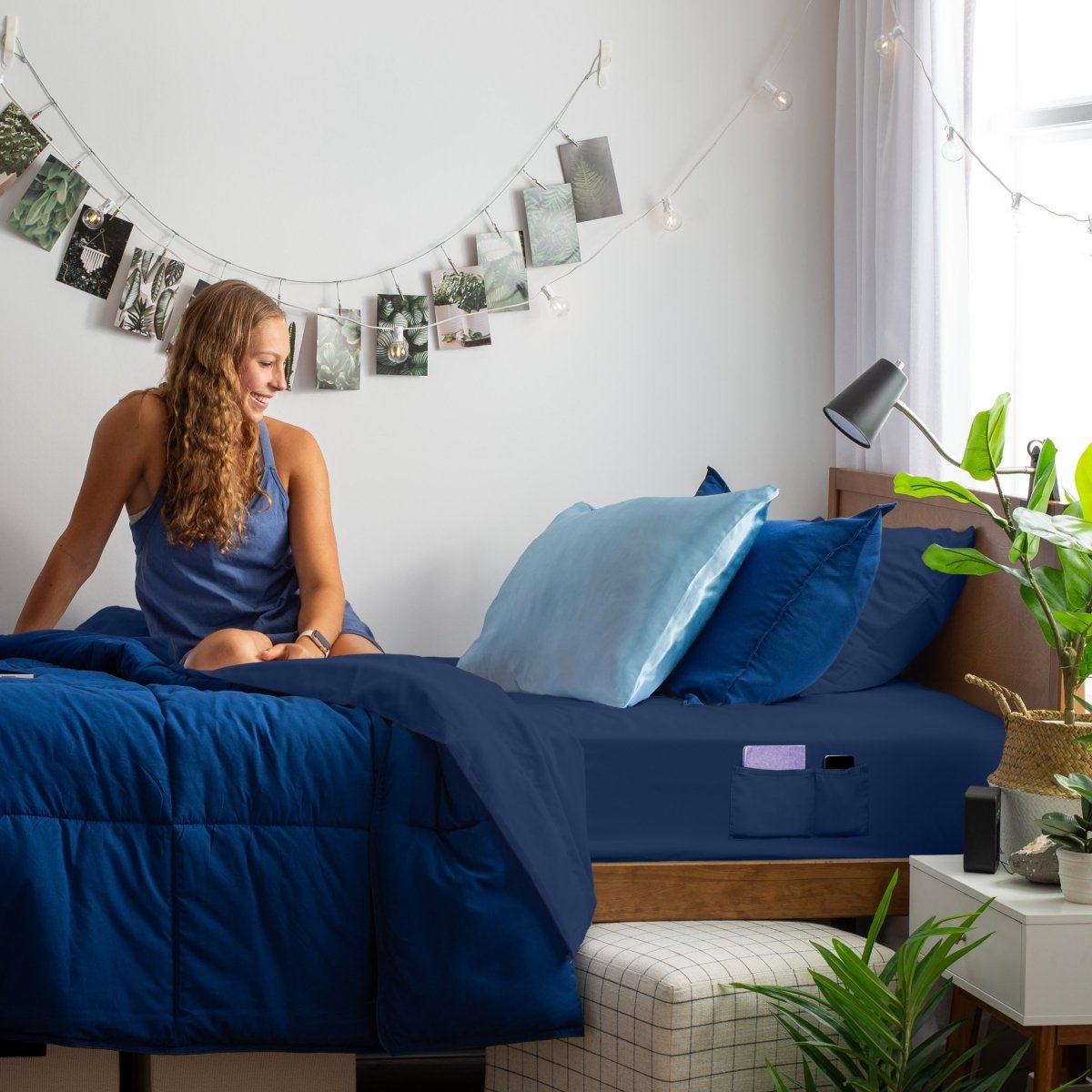 Woman sitting on a bed with blue bedding, surrounded by plants and decor.