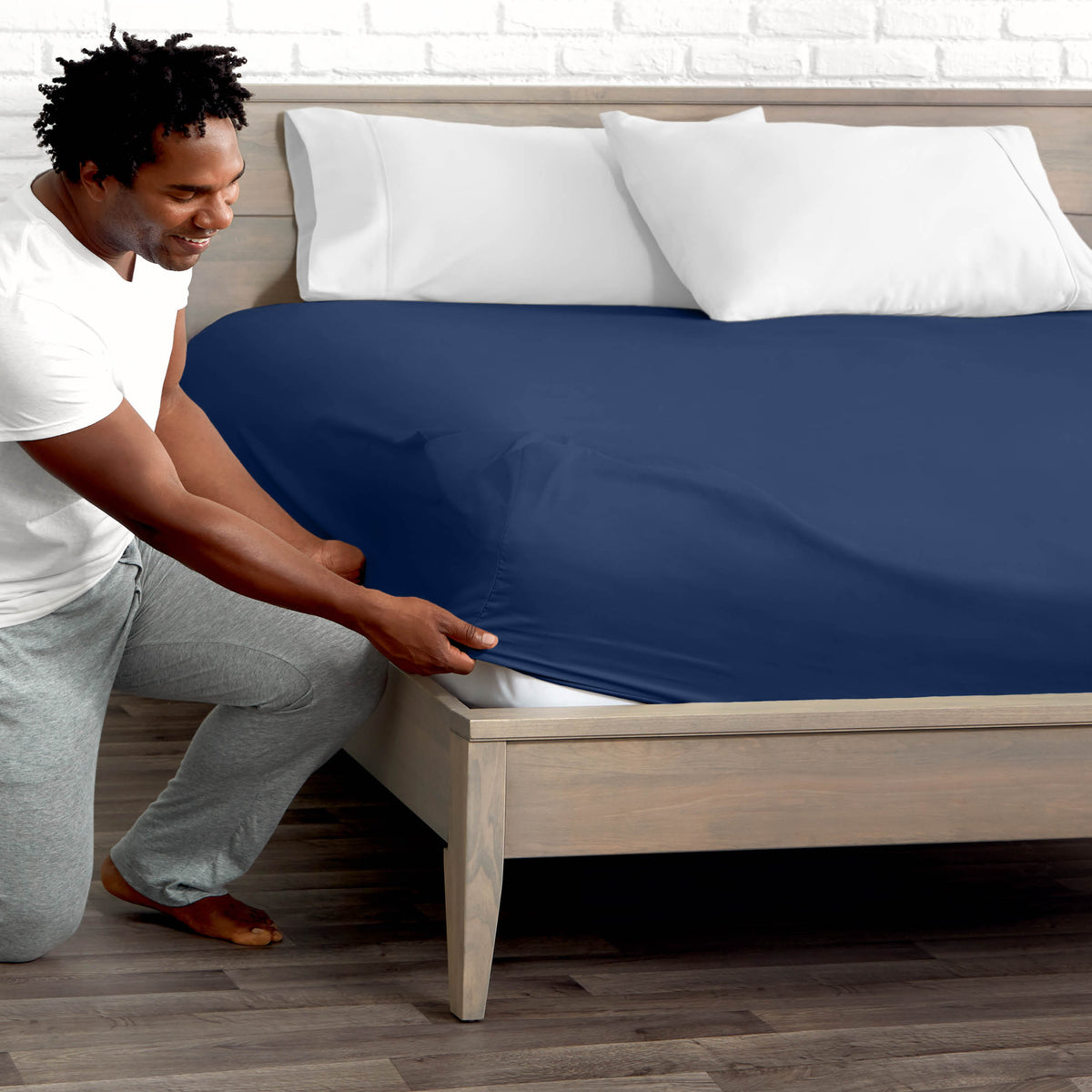 Man adjusting fitted sheet on a bed with white pillows.