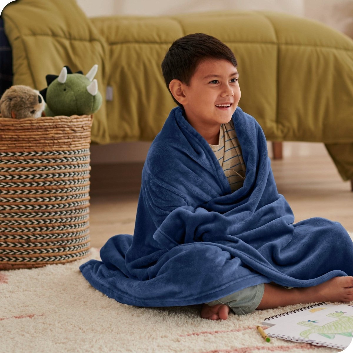A boy is sitting on the floor of his bedroom with a microplush blanket wrapped around him