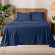 Organic cotton jersey sheets on a bed with a wicker headboard in a room with plants and a lamp.