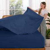 Woman putting sheet on a bed in a bright room with large windows.