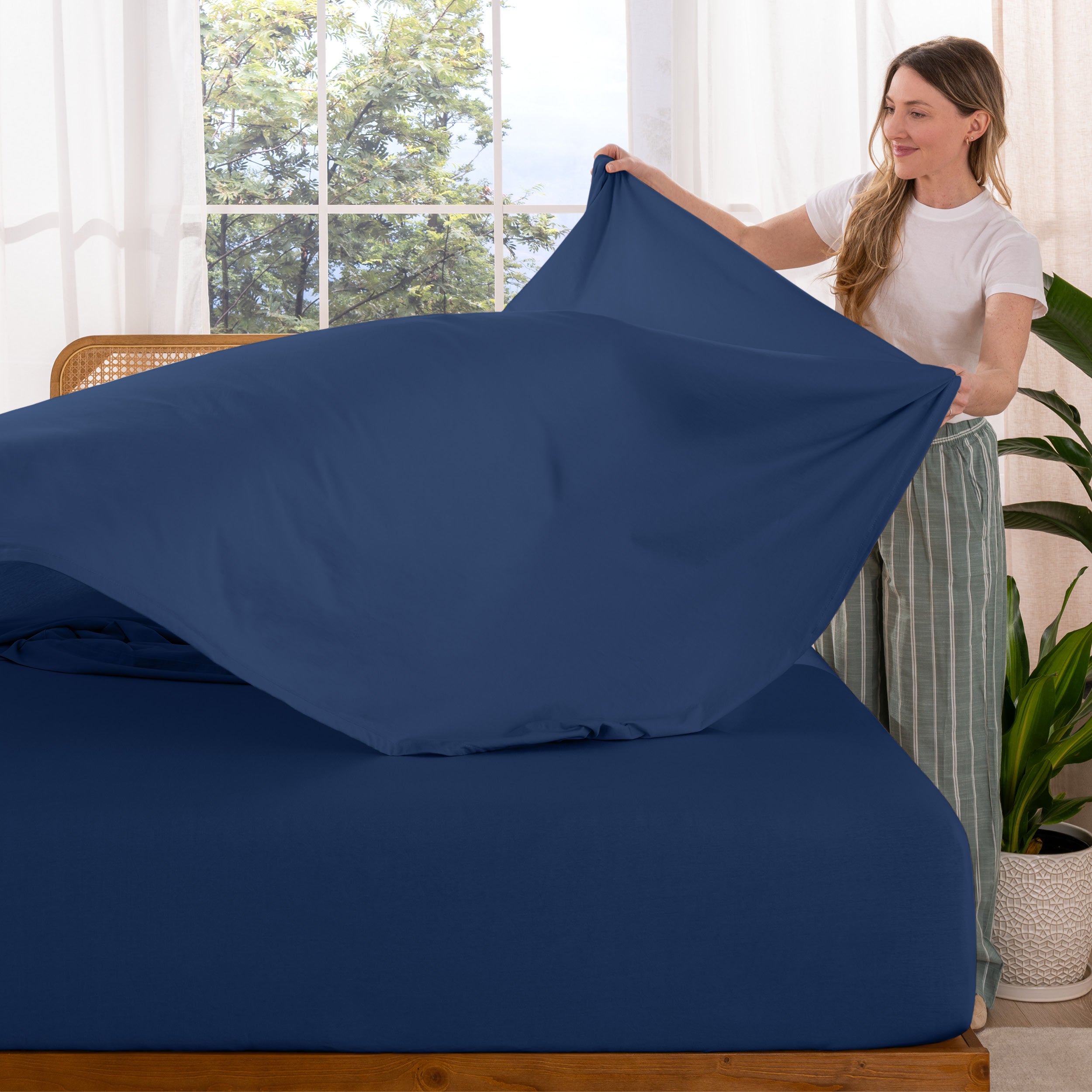 Woman putting sheet on a bed in a bright room with large windows.
